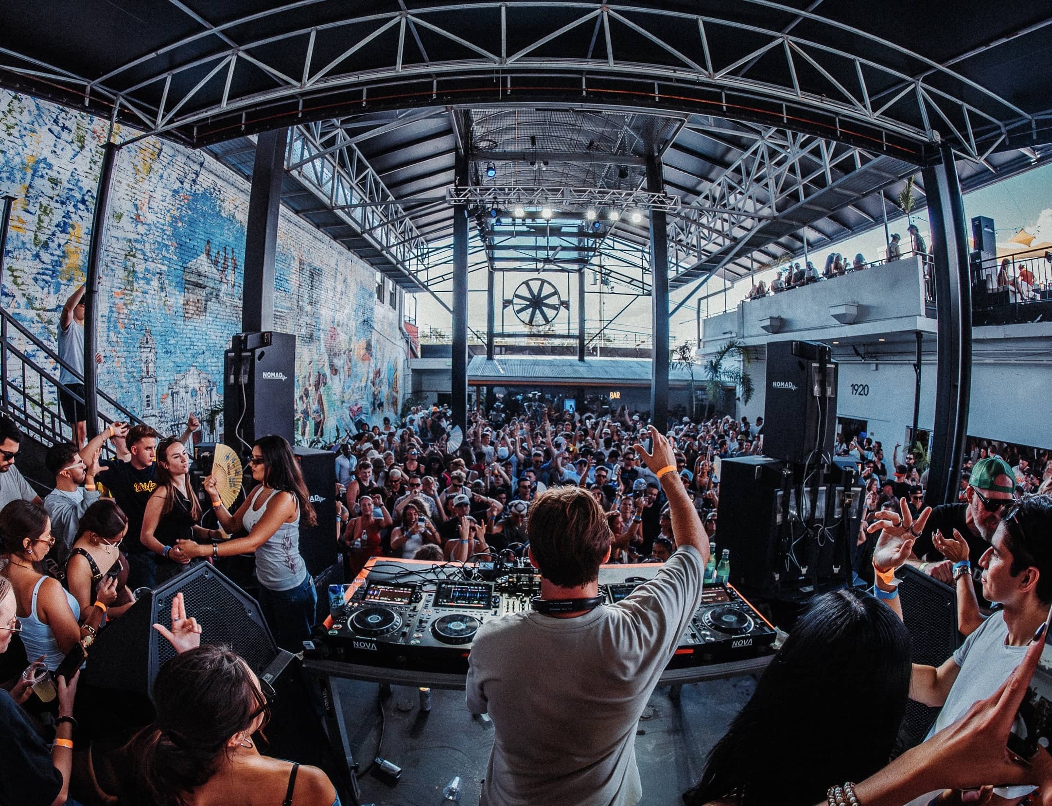 View from the stage across a packed open-air Miami warehouse: crowd, mezzanine and roof trusses in daylight